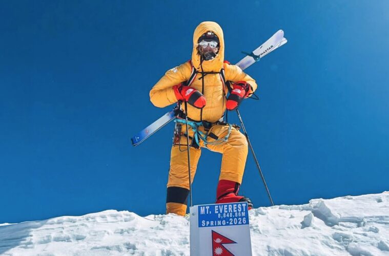 Mountaineer in orange suit standing on snow, holding skis, with Mt. Everest sign in the foreground against a clear blue sky.