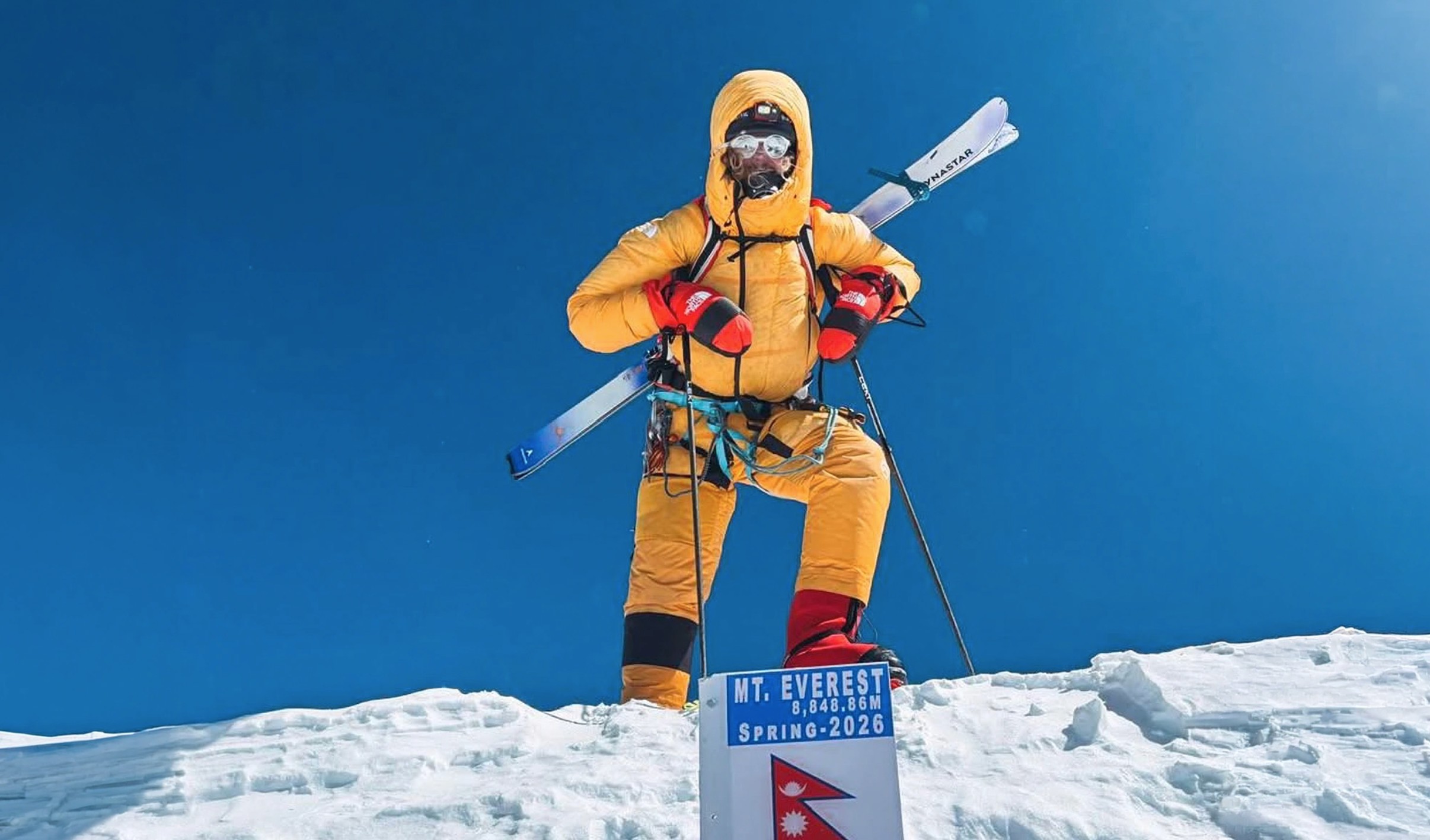 Mountaineer in orange suit standing on snow, holding skis, with Mt. Everest sign in the foreground against a clear blue sky.
