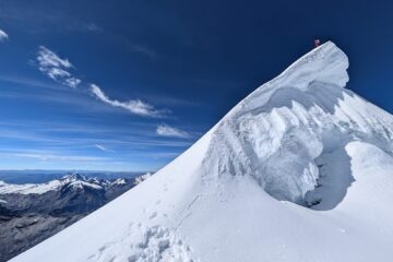 A climber stands at the summit of a sharp snow ridge, high above a rugged mountain range under a clear blue sky.