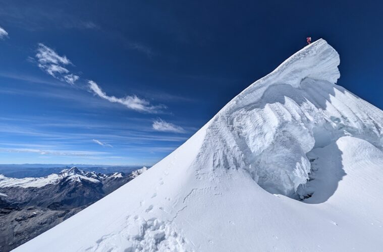 A climber stands at the summit of a sharp snow ridge, high above a rugged mountain range under a clear blue sky.