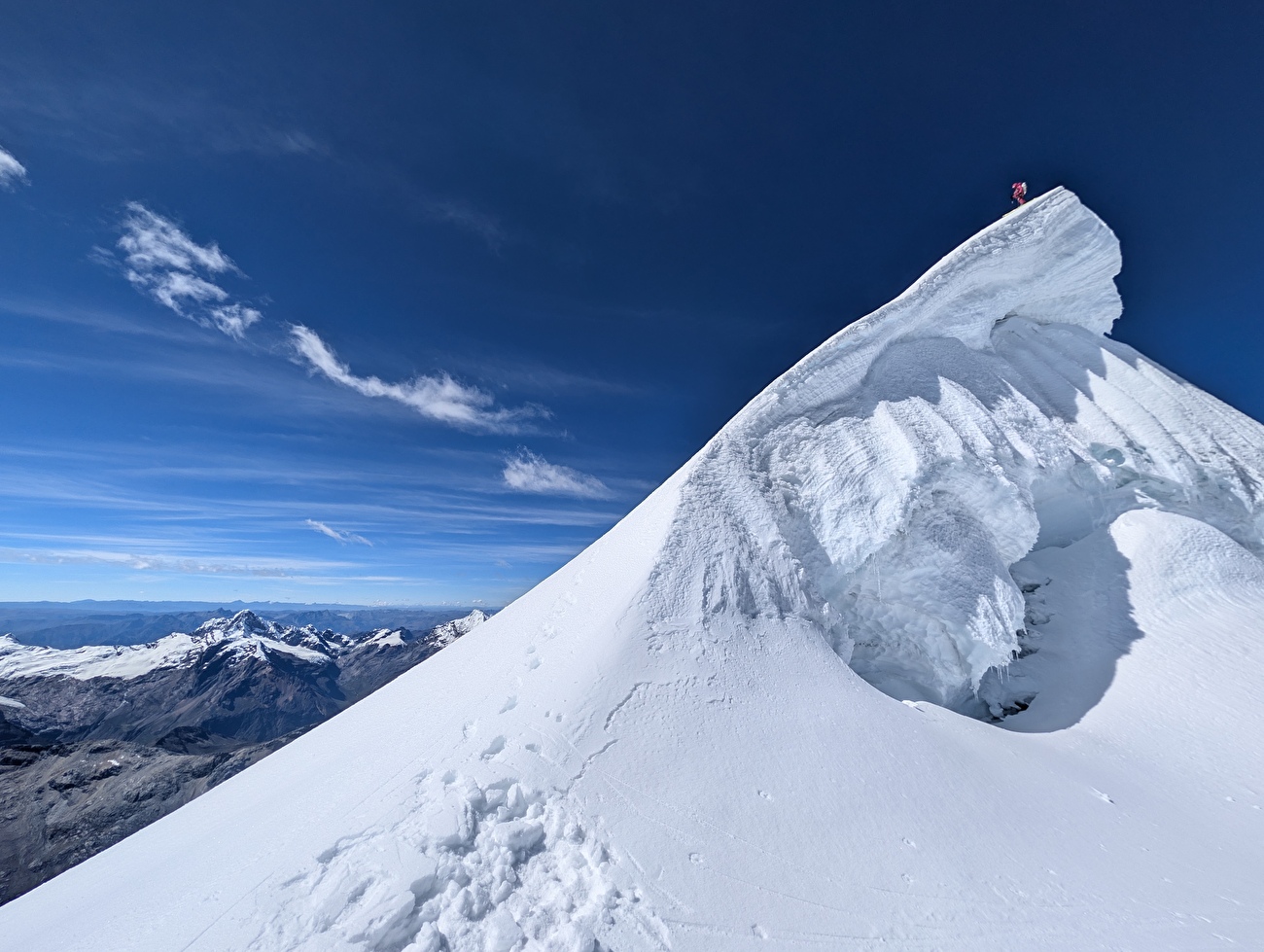 A climber stands at the summit of a sharp snow ridge, high above a rugged mountain range under a clear blue sky.